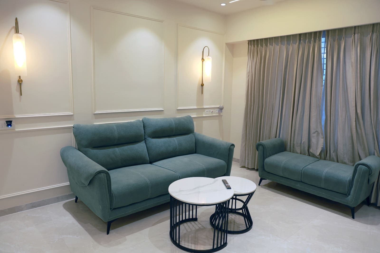 Living room with sage sofas, a marble-top nesting table, wall sconces and sheer curtains